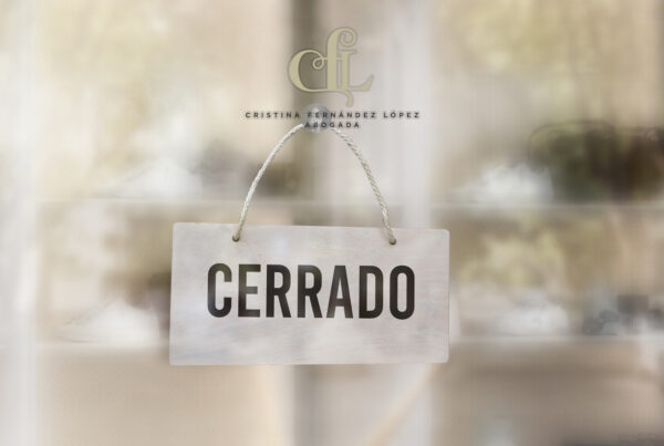 'CERRADO' sign hanging on a glass storefront door with a blurred shop interior in the background; logo above.