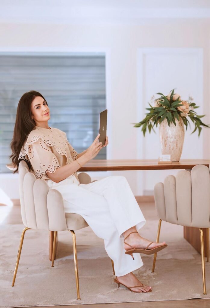Woman with long dark hair sits at a wooden dining table, holding a tablet for a selfie in a bright, elegant room.