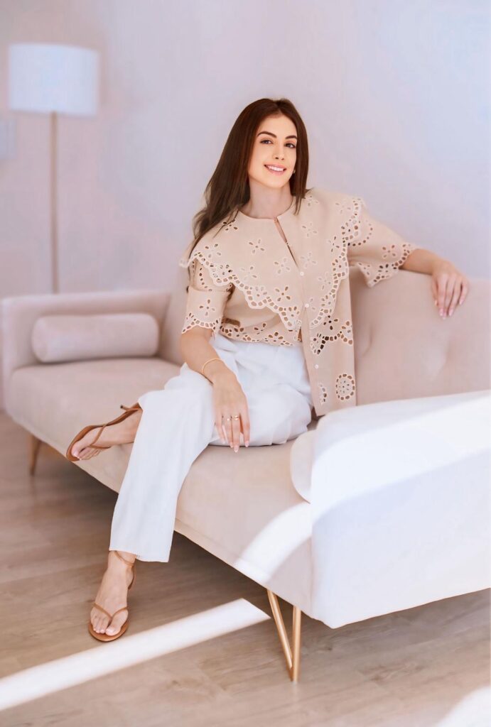 Smiling woman with long brown hair sits on a beige sofa in a light, airy room, wearing a beige eyelet blouse and white trousers with tan sandals.