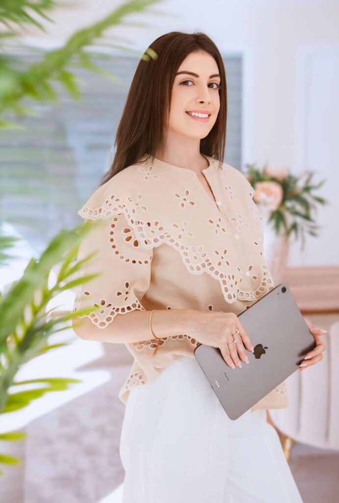 Smiling woman in a beige embroidered blouse holds a silver tablet indoors with greenery in the foreground.