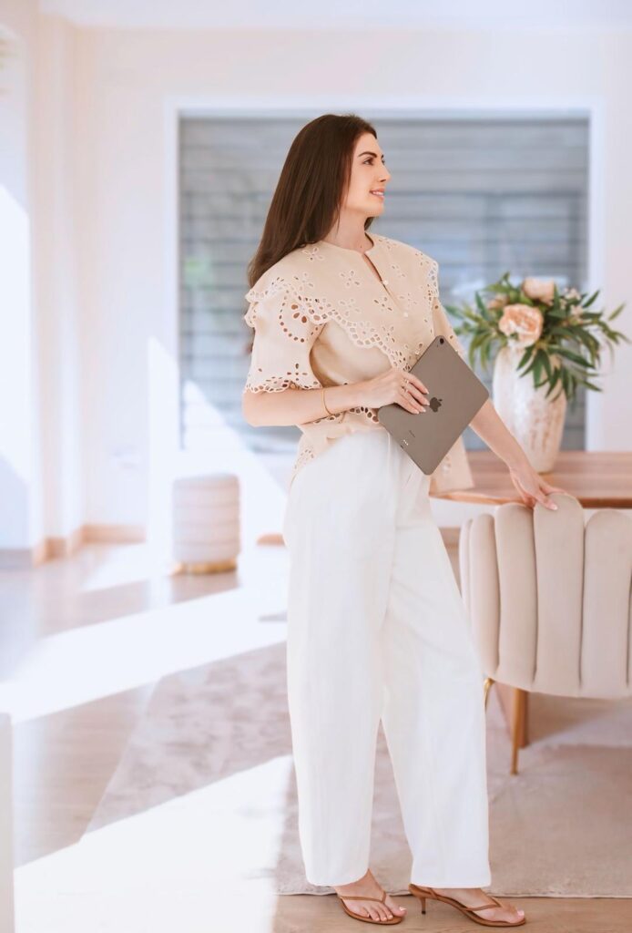 A woman in a beige eyelet blouse and white wide-leg pants walks through a bright, modern living space while holding a gray tablet.